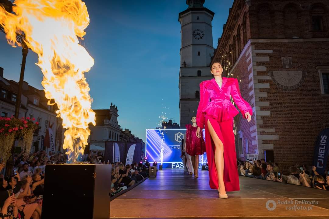 Modelki w czerwonych sukniach na tle ratusza w Sandomierzu i ekranu LED, na którym wyświetlane jest logo projektanta